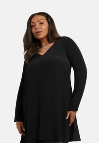 Svart långärmad tunika med V-ringning, tillverkad av ett mjukt tyg, med sidokilar och en avslappnad passform. Accenterad med kedjesmycken.