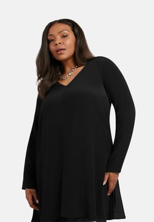 Tunique noire à manches longues avec un décolleté en V, fabriquée à partir d'un tissu lisse, dotée d'une fente latérale et d'une coupe décontractée. Accentuée avec des bijoux en chaîne.