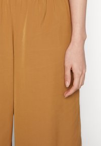 Tan, lette stofbukser med elastisk talje. Teksturen ser glat ud med en subtil glans. En hånd hviler afslappet på siden.