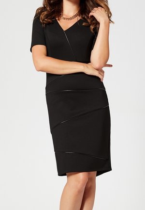 Mujer que lleva un vestido negro ajustado con mangas cortas y un ribete brillante en diagonal, con los brazos cruzados y un collar de cadena dorada.