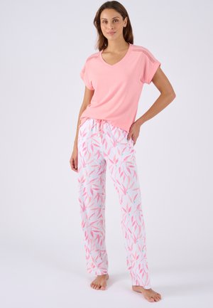 Mujer de pie descalza con una camiseta rosa de manga corta y pantalones de pijama blancos con motivos de hojas rosas sobre fondo blanco.