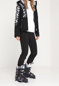 Czarna kurtka narciarska z wzorem pepitki na ramionach, w połączeniu z czarnymi spodniami i czarnymi butami narciarskimi z fioletowymi akcentami i srebrnymi elementami.