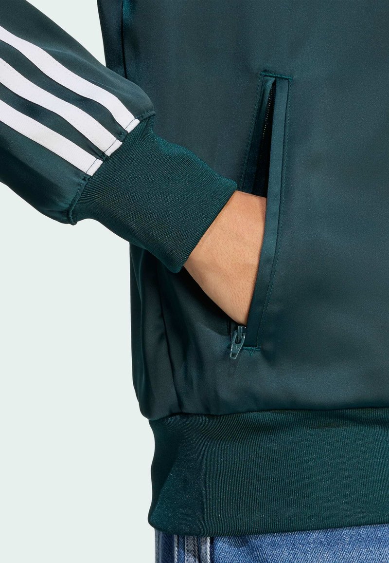 Grøn satinjakke med hvide striber på ærmerne, lynlåslomme, ribstrikkede manchetter og kant. Glat tekstur, afslappet design.