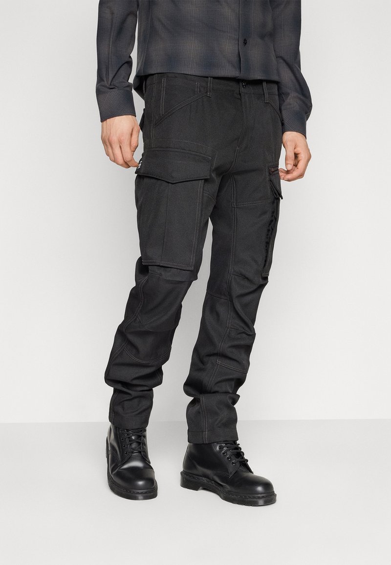 Homme portant un pantalon cargo noir avec plusieurs poches et des bottes noires à lacets, debout devant un fond uni.