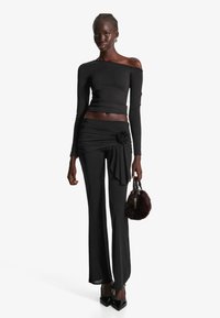 Svart långärmad off-the-shoulder topp som matchas med högmidjade plisserade byxor med blommiga accenter; håller en liten svart fluffig väska.