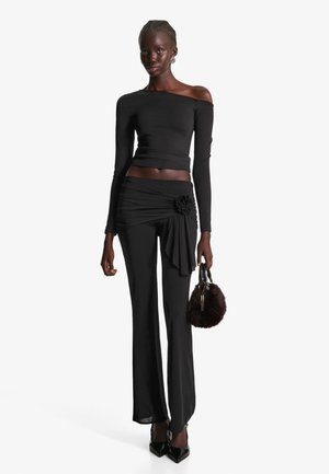 Fekete, hosszú ujjú, vállon kívüli felső, magas derekú, plisszírozott nadrággal párosítva, virágos akcentussal; egy kis fekete, bolyhossá tett táskát tart a kezében.