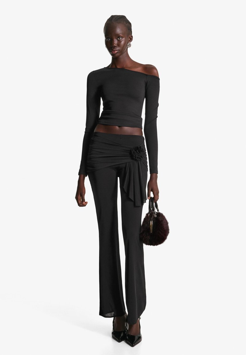 Svart långärmad off-the-shoulder topp som matchas med högmidjade plisserade byxor med blommiga accenter; håller en liten svart fluffig väska.