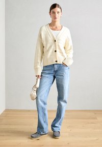 Cardigan a coste crema con due bottoni, abbinato a jeans dritti azzurri e scarpe grigie, mentre tiene una borsa beige.