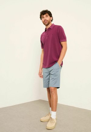 Muž s kučeravými vlasmi, ktorý má na sebe tmavofialové polo tričko, svetlomodré šortky, béžové tenisky a biele ponožky, stojí pri obyčajnej stene.