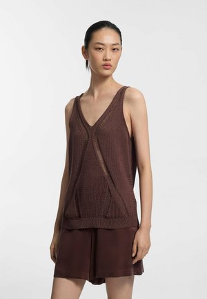 Femme portant un haut marron sans manches à col en V avec des motifs texturés et un short marron ample, debout devant un fond clair uni.
