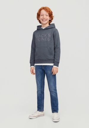 Garçon aux cheveux roux souriant, portant un sweat à capuche gris foncé avec « JACK & JONES », un jean bleu et des baskets blanches, debout devant un fond clair uni.