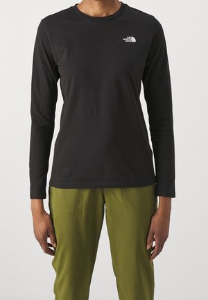 Person iført sort langærmet skjorte med hvidt The North Face-logo og olivengrønne bukser, stående mod en ensfarvet lys baggrund.