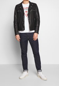 Chaqueta de cuero negra estilo motero con bolsillos con cremallera, camiseta gráfica blanca, jeans de mezclilla oscuros y zapatillas blancas. Conjunto casual con texturas superpuestas.