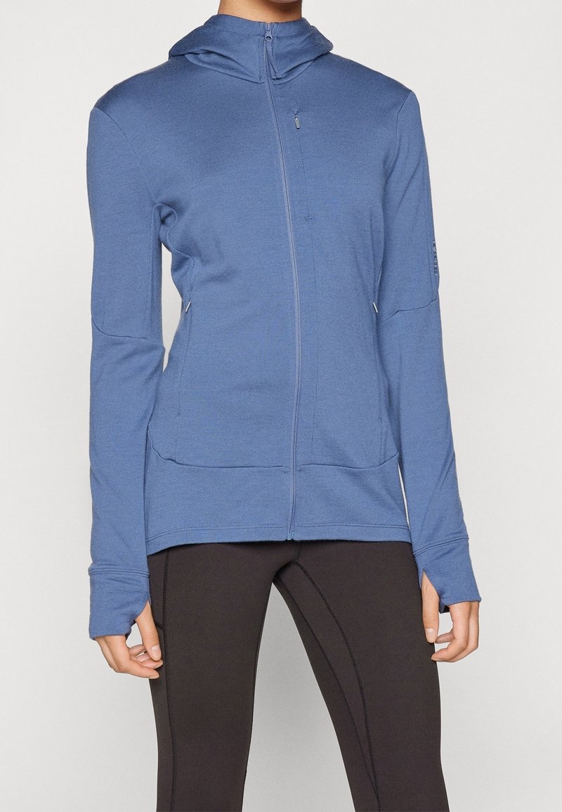 Personne portant un sweat à capuche bleu zippé avec des trous pour les pouces et un legging noir ajusté sur un fond uni.