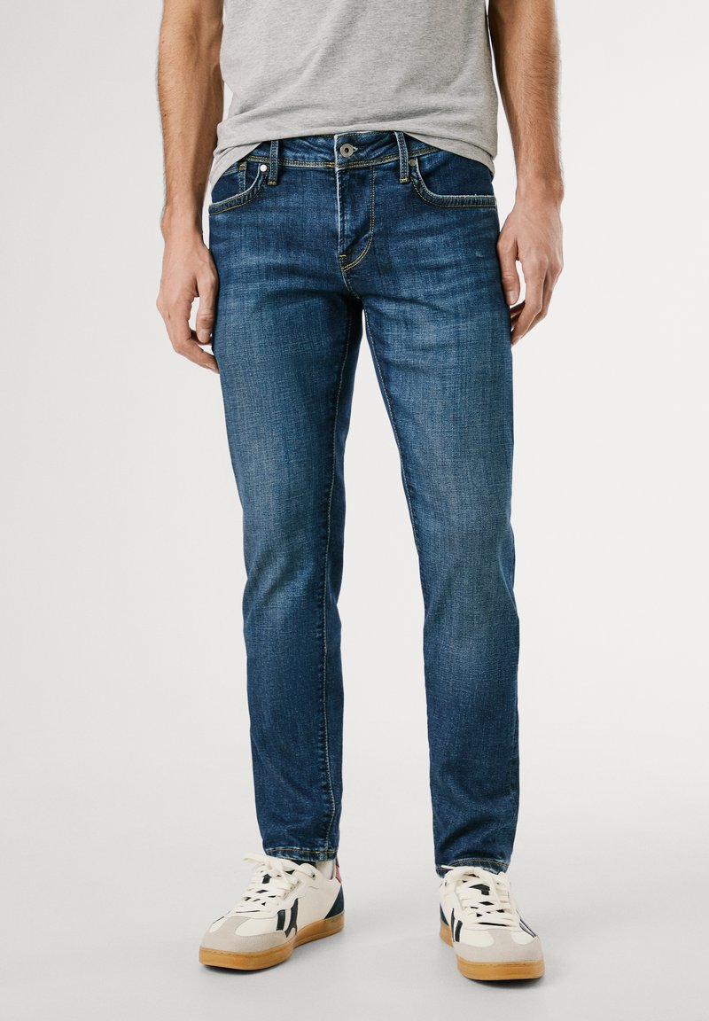 Blauwe denim jeans met een slanke pasvorm, voorzien van een klassieke vijf-pocketontwerp, gele stiksels en een knoopsluiting. Draagt met witte sneakers.
