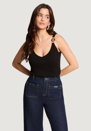 Donna con lunghi capelli castani, che indossa un top nero senza maniche con dettaglio ad anello dorato sulla spalla e jeans blu scuro a vita alta.