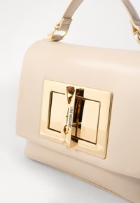 Beige syntetisk håndveske med en glatt tekstur, som har en fremtredende gulllås og branding på forsiden, supplert med en strukturert form.