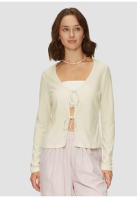 Hellecremiger, gerippter Cardigan mit tiefem V-Ausschnitt, Bindeband-Frontverschluss und langen Ärmeln, über einem weißen Oberteil und rosa gestreiften Hosen getragen.
