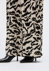 Εκρού και μαύροι παντελόνες με αφηρημένο σχέδιο και φαρδιά γραμμή, συνδυασμένες με μυτερές μαύρες δερμάτινες μπότες με ψηλό τακούνι, σε λευκό φόντο.