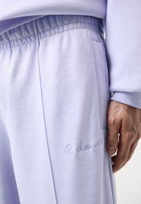 Világos lila melegítőnadrág, rugalmas derékkal, oldalsó zsebekkel és hímzett "R. Lacoste" logóval a lábon. Sima pamutkeverék anyag.