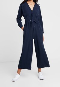 Mujer vestida con un mono de pernera ancha de manga larga en azul marino, con cintura ajustable y zapatillas blancas, de pie con las manos en los bolsillos.