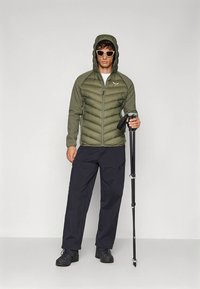 Padded olive green jacket with hood, zip closure, and contrasting sleeves, paired with dark pants and black boots. Holding a trekking pole.