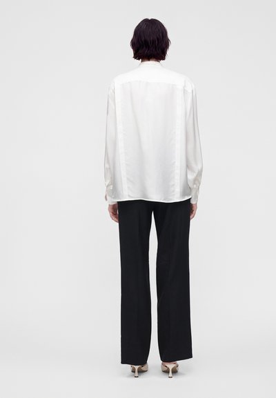 Personne vue de dos portant un chemisier blanc à manches longues, un pantalon noir à jambes larges et des chaussures à talons hauts beiges sur un fond uni.