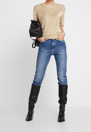 Mujer con top de punto dorado, jeans azules y botas negras hasta la rodilla, llevando una mochila de cuero negro sobre un hombro.