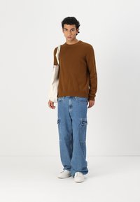 Pull marron, jean cargo bleu ample avec poches avant, baskets blanches et sac fourre-tout crème. Design simple avec une coupe décontractée.