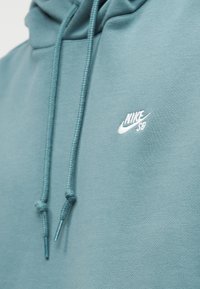 Sweat à capuche Nike SB de couleur bleu sarcelle avec cordons apparents et petit logo blanc brodé sur la poitrine.