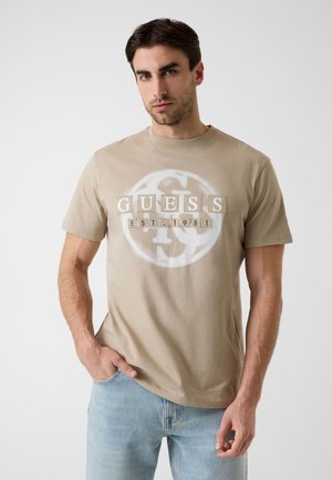 Mann trägt ein beigefarbenes GUESS-T-Shirt mit dem Schriftzug "GUESS EST. 1981" und hellblaue Jeans, steht mit einer Hand in der Tasche vor weißem Hintergrund.