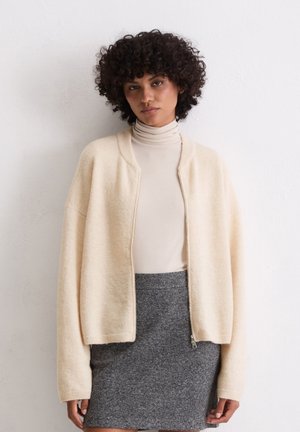 Cardigan in maglia morbida color beige chiaro, con zip frontale e maniche ampie, abbinato a un dolcevita aderente e a una minigonna grigia. Texture complessivamente liscia.