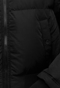 Μαύρο μαξιλαρωτό μπουφάν σε κοντινή λήψη που δείχνει φερμουάρ, τσεπάκι με κουμπί και λεπτομέρειες στα μανίκια.