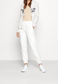 Světle šedá mikina na zip s námořnickým písmem, béžový přiléhavý top, bílé joggingové kalhoty s elastickými manžetami a bílé tenisky se zelenými akcenty.