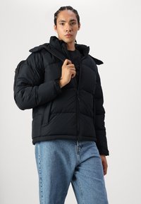 Sort polstret jakke med lynlås foran og hætte, med en glat tekstur og ribbede manchetter, matchende lyseblå denimbukser.
