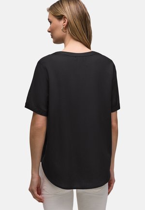 Kobieta o jasnobrązowych włosach, ubrana w luźną czarną bluzkę na krótki rękaw i białe spodnie, widziana od tyłu na jednolitym białym tle.