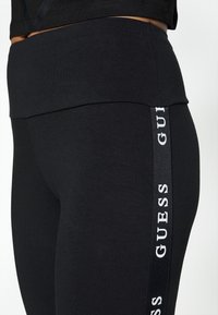 Černé legíny s širokým pasem, na vnější straně s lesklým vertikálním logem. Hladká, elastická textura materiálu.