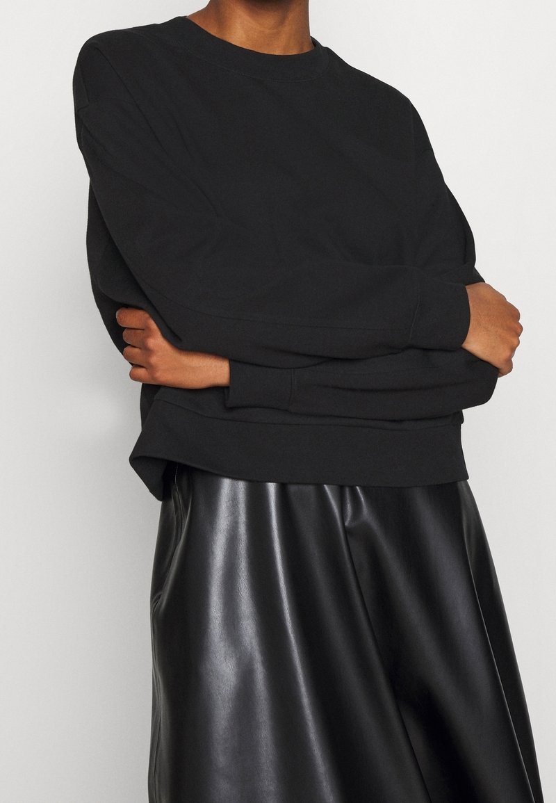 Sweat-shirt en coton noir avec un col rond et des manches longues, associé à une jupe noire en fausse cuir taille haute. Texture lisse.