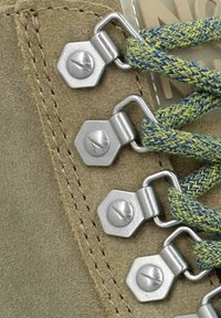 Gros plan sur les œillets en daim beige d'une botte avec des crochets hexagonaux argentés tenant des lacets tissés bleus et jaunes, lacés serrés.