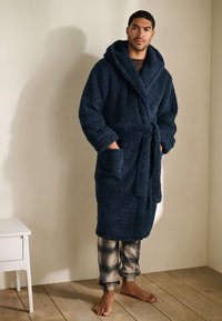 Marineblå, blød badekåbe med hætte, som har et bælte og to forlommer. Båret over mønstrede ternede pyjamasbukser. Trægulv.