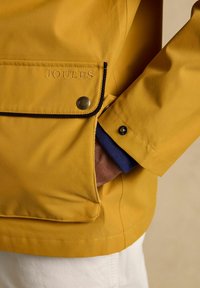 Hand in de zak van een mosterdgele jas met een gerafelde klepzak met het borduursel "JOULES" en zwarte bies, gedragen over een marineblauwe mouw.