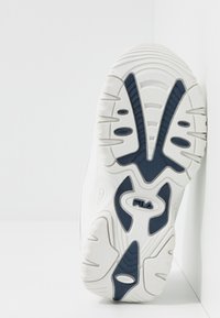 Semelle blanche de sneaker avec des sections de grip en caoutchouc bleu marine et le logo Fila au centre, présentée sur un fond blanc.