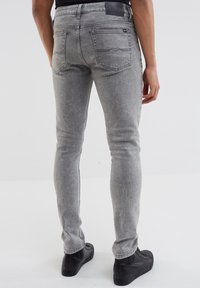 Jean en denim gris à coupe slim, avec poches arrières, coutures minimales et étiquette de ceinture noire, assortis à des chaussures noires.