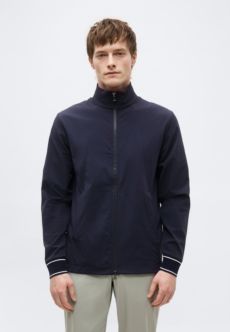 Νέος άνδρας που φοράει μπλε ζακέτα με φερμουάρ και ψηλό γιακά, με ανοιχτόχρωμο παντελόνι, στέκεται μπροστά σε λευκό φόντο.