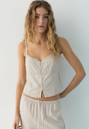 Femeie blondă cu păr ondulat, purtând un top crem cu nasturi și pantaloni largi asortați, pe un fundal simplu, deschis la culoare.