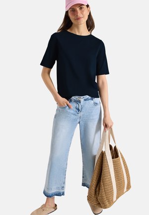 Kvinde iført en sort top med korte ærmer, lyse korte jeans, lyserød kasket, beige slip-on sko, der holder en stor vævet taske med brede stropper.