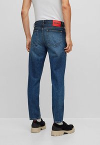 Blå denimjeans med rak passform, med en röd logotypapplikation på midjan och två bakfickor. Bärs med svarta sneakers.