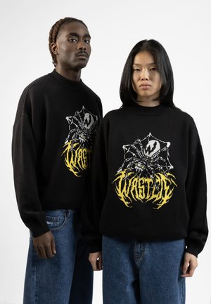 Black knit sweaters featuring a spider graphic in grey and yellow, paired with blue denim jeans. Sweaters have a relaxed fit and high neck.