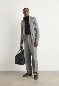 Grå zip-up cardigan över en svart polo, i kombination med texturerade grå byxor. En svart duffelväska och mörka skor fulländar outfiten.