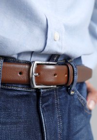 Brown leather belt with a silver metal rectangular buckle, worn over blue denim jeans and a light blue shirt. Stitching is visible on the jeans.